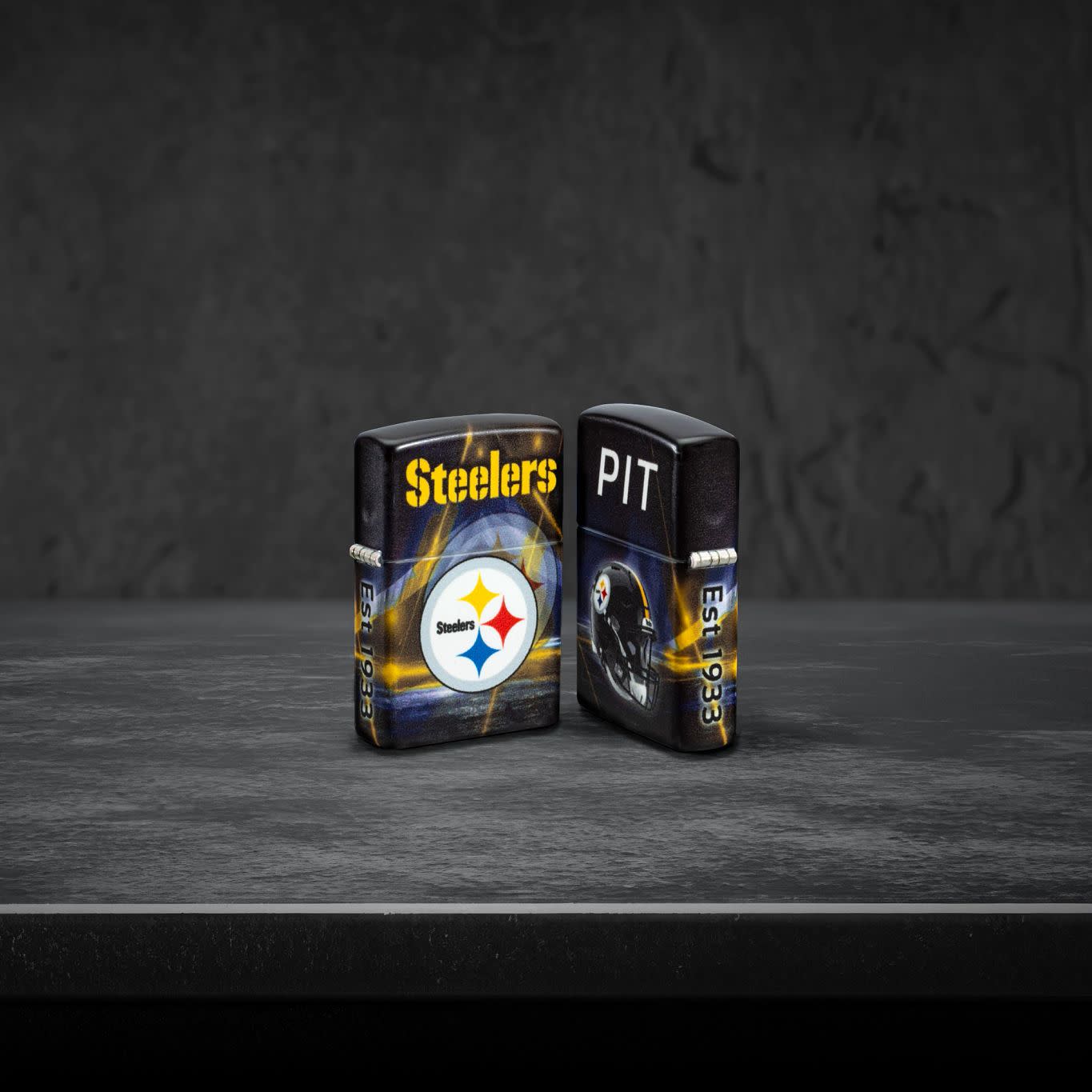 Lifestyle image of two Zippo NFL Pittsburgh Steelers 540 Matte Windproof Lighters, one showing the front of the lighter and the other showing the back, standing on a dark grey surface.