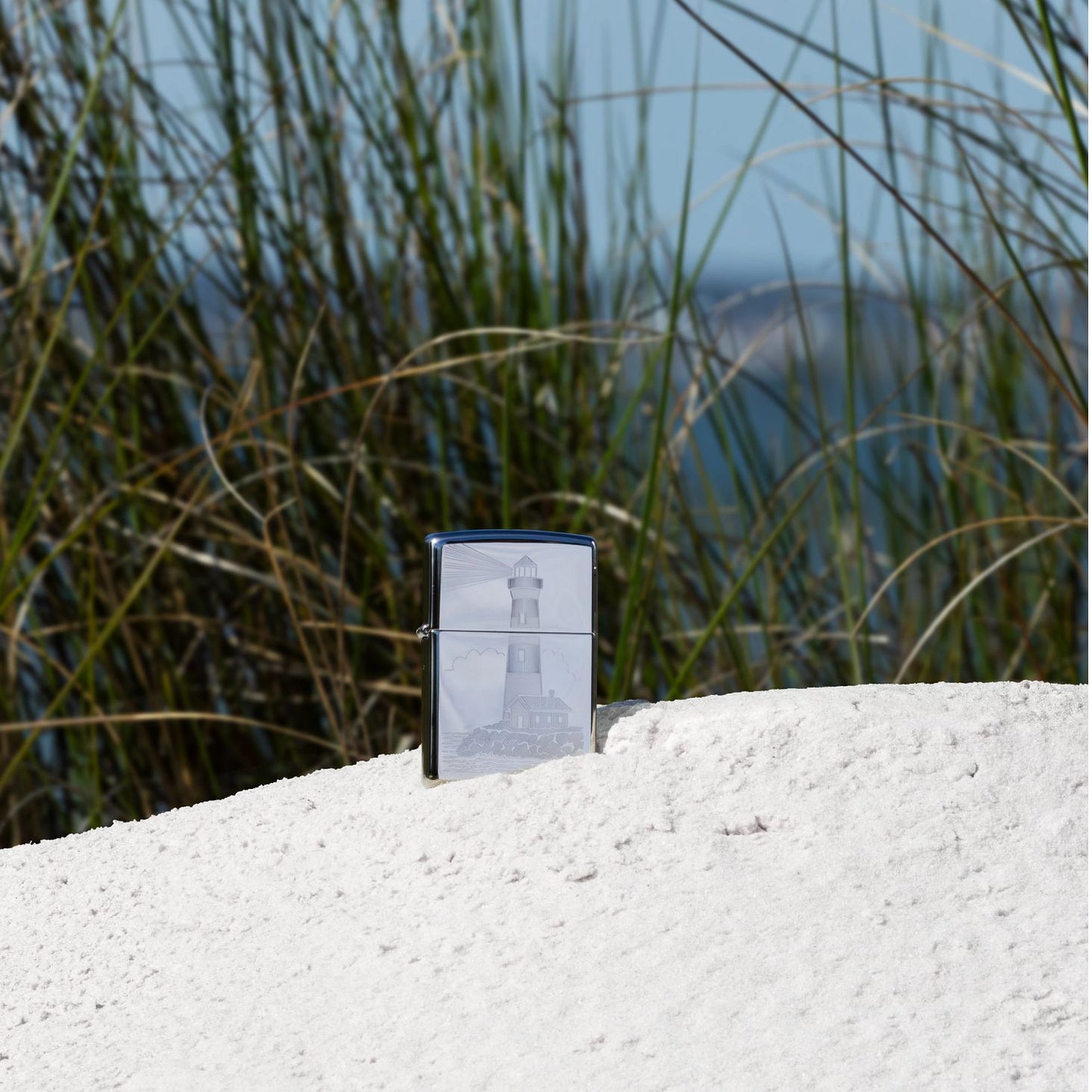 Lifestyle image of Zippo Lighthouse Design High Polish Chrome Windproof Lighter standing in the sand with seagrass in the background.