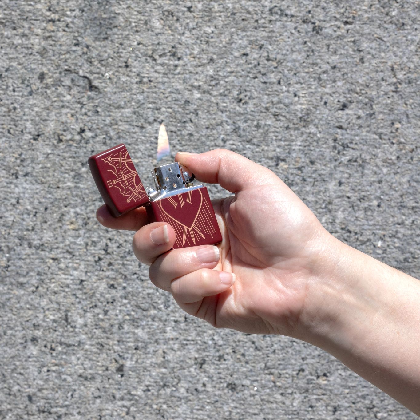 Lifestyle image of Zippo Three of Swords Design Merlot Windproof Lighter against a gray marbled background, being lit in hand.