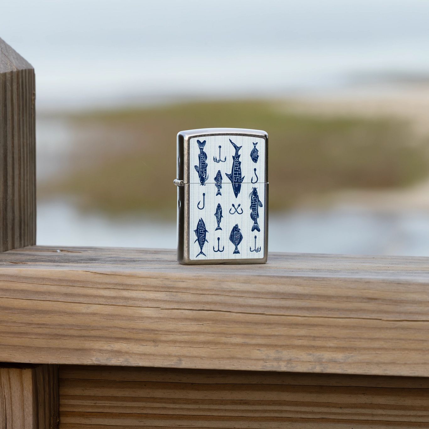 Lifestyle image of Zippo Off the Hook Design Street Chrome Windproof Lighter standing on a wooden railing with a blurred seascape in the background.