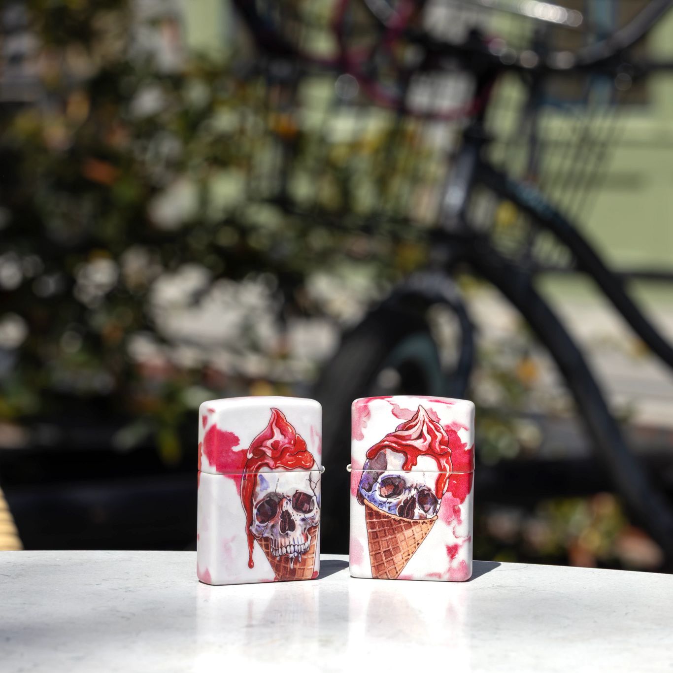 Lifestyle image of two Zippo I Scream Cone Design 540 Matte Windproof Lighters, one showing the front of the lighter and the other showing the back, standing on an outdoor table with a blurred scene behind it.