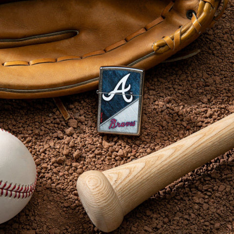 Lifestyle image of MLB® Atlanta Braves™ Street Chrome™ Windproof Lighter laying on a baseball field with a glove, ball, and bat.