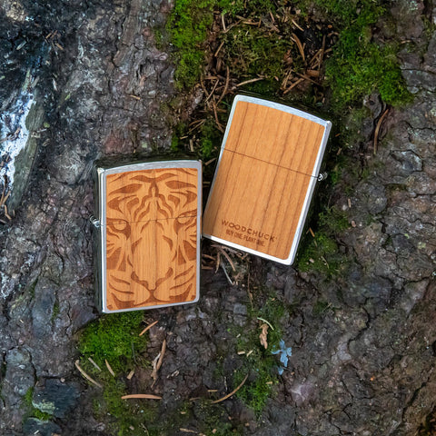 Lifestyle image of two WOODCHUCK USA Cherry Tiger Head Emblem Windproof Lighters laying on a log. One lighter is showing the front of the design with the other showing the back.