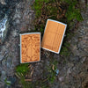 Lifestyle image of two WOODCHUCK USA Cherry Tiger Head Emblem Windproof Lighters laying on a log. One lighter is showing the front of the design with the other showing the back.