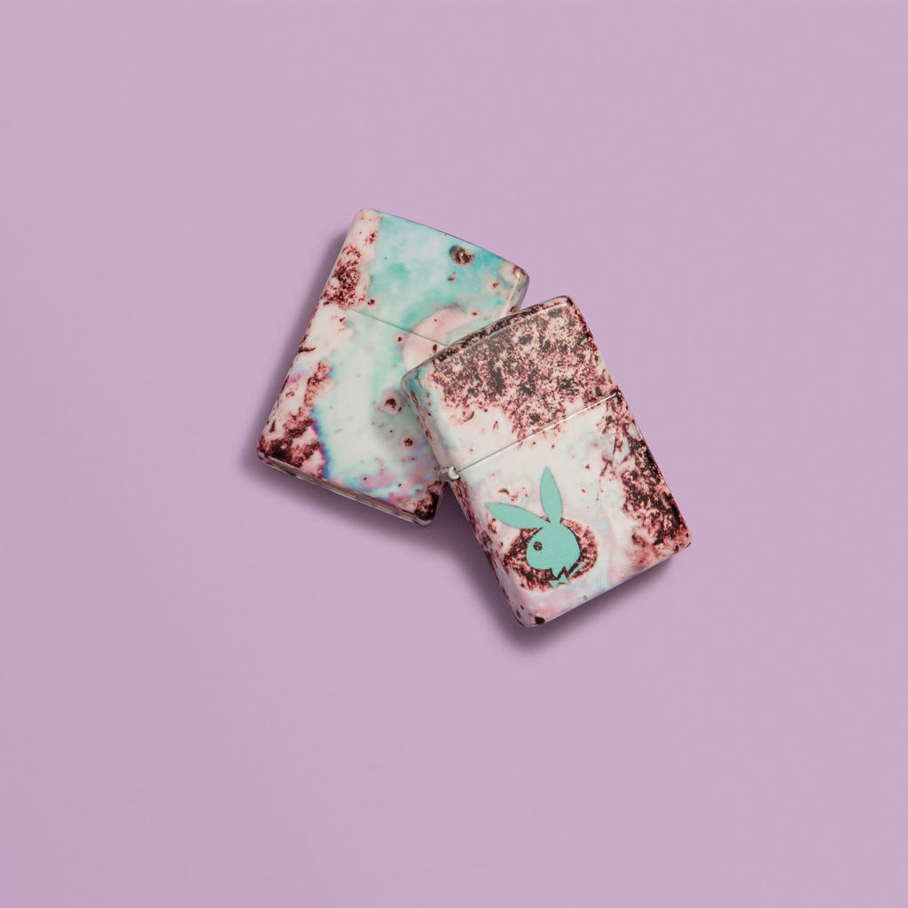 Lifestyle image of two Playboy Teal 540 Color Design Windproof Lighters, laying on a pink background.