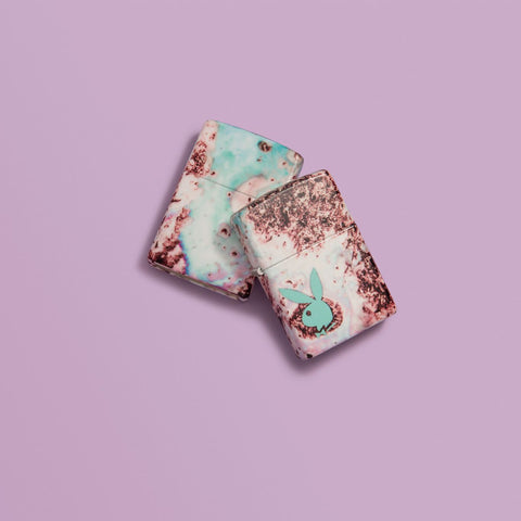Lifestyle image of two Playboy Teal 540 Color Design Windproof Lighters, laying on a pink background.
