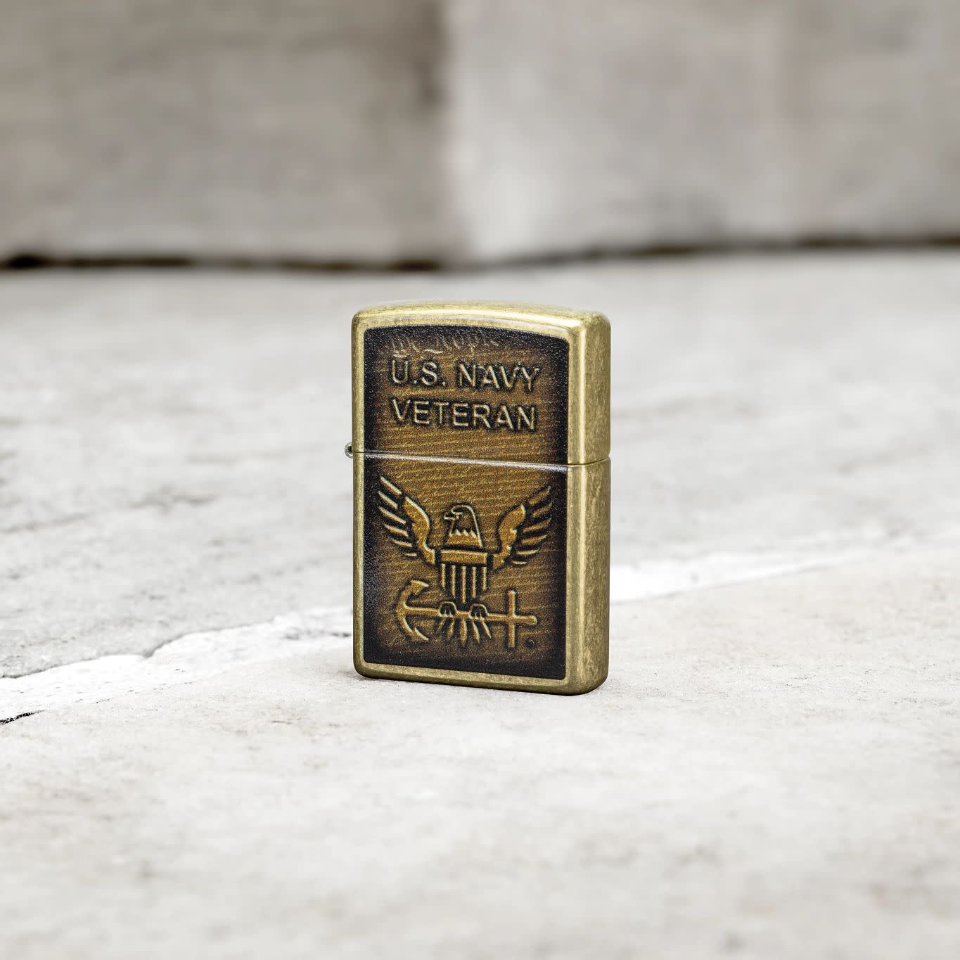Lifestyle image of Zippo United States NavyÂ® Street Brass Windproof Lighter standing on a grey tile floor.