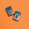 Lifestyle image of two Harley-Davidson 360° Flames High Polish Blue Windproof Lighters laying on an orange background. One lighter is showing the front, while the other is showing the back.