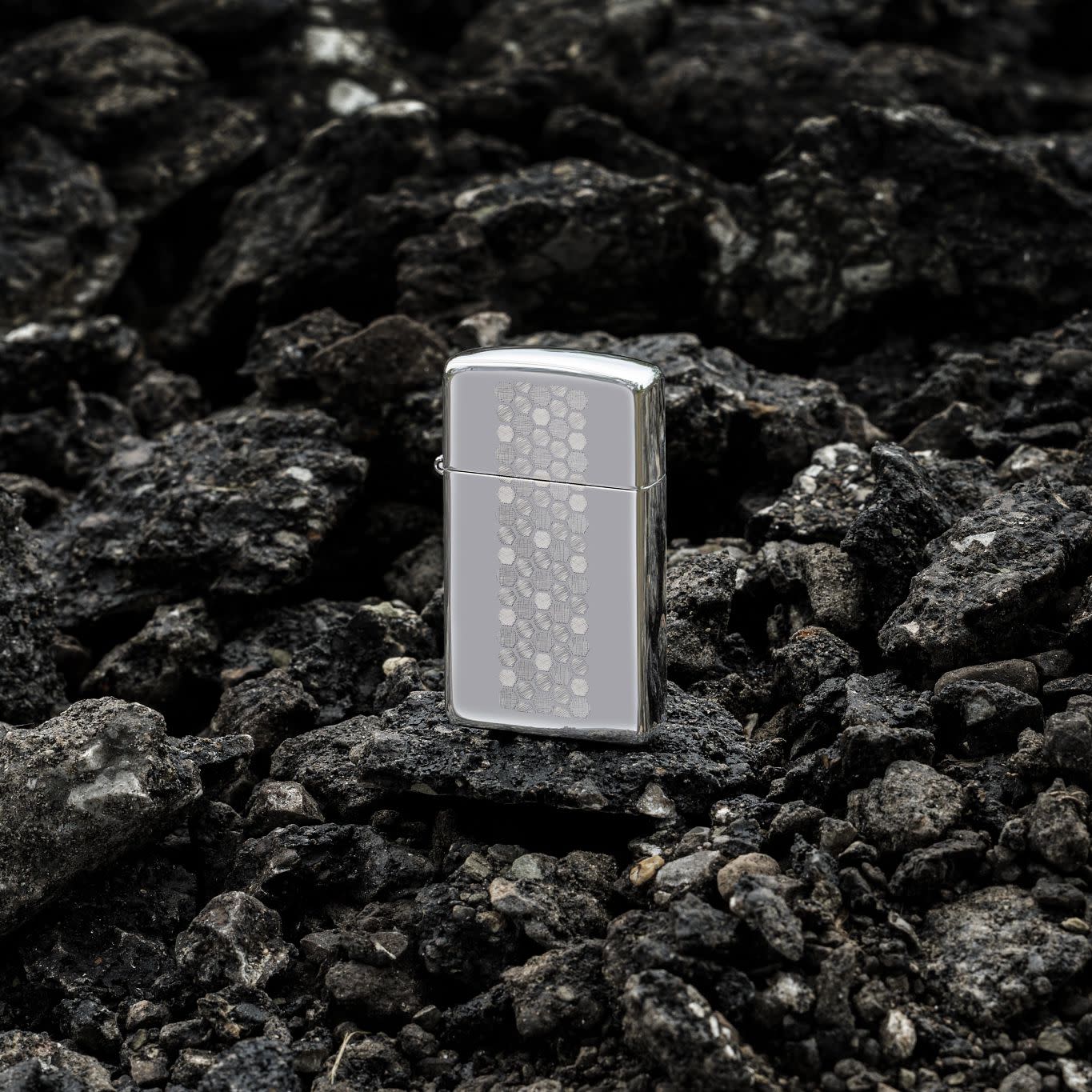 Lifestyle image of Zippo SlimÂ® Honeycomb Design High Polish Chrome Windproof Lighter standing on dark, loose rocks.