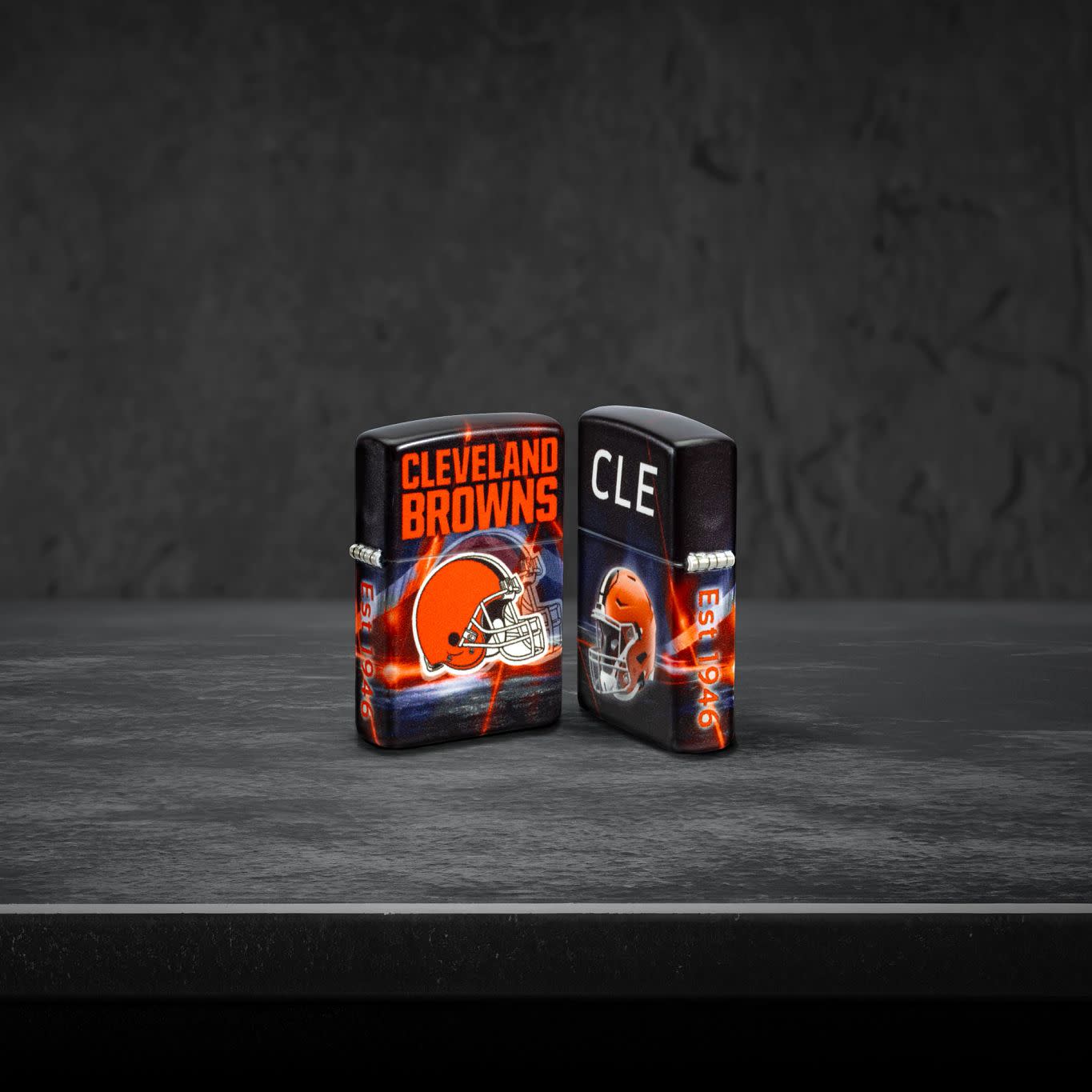 Lifestyle image of two Zippo NFL Cleveland Browns 540 Matte Windproof Lighters, one showing the front of the lighter and the other showing the back, standing on a dark grey surface.