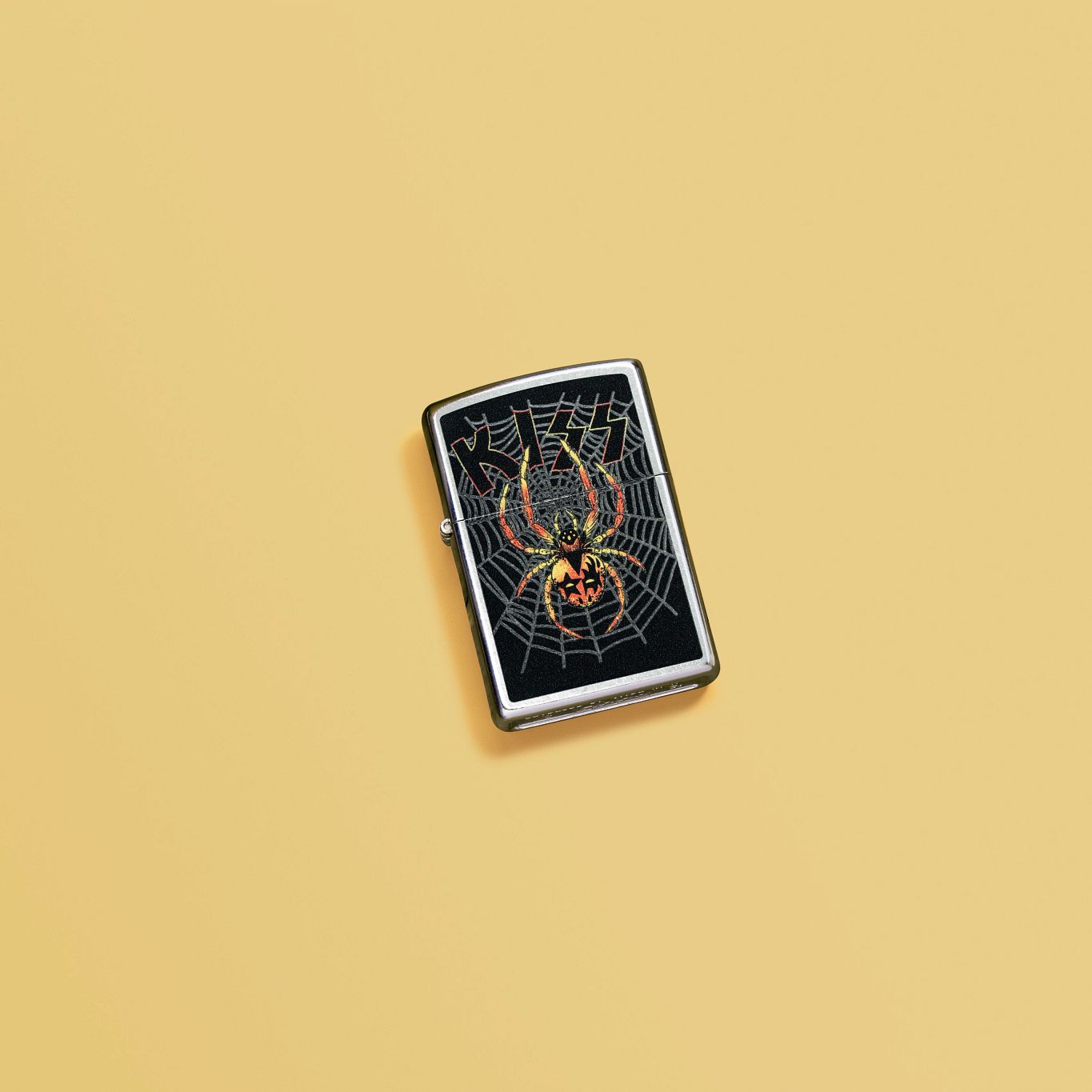 Lifestyle image of Zippo KISS® Spider Design Street Chrome Windproof Lighter laying on a pale-yellow surface.