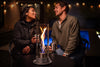 Lifestyle image of the Zippo FlameScapes™ Spiral Fire Feature XL in black, lit on a table with a cityscape and couple in the background.