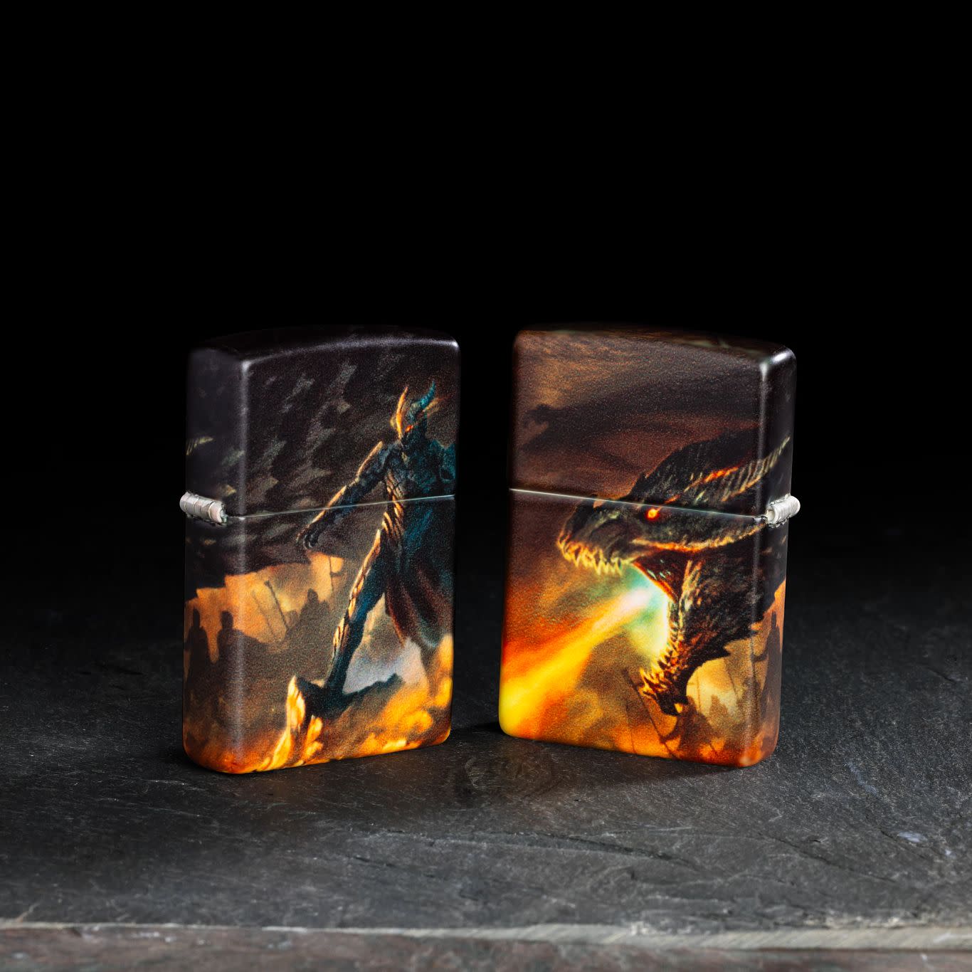 Lifestyle image of two Zippo Knight and Dragon Design 540 Matte Windproof Lighters, one showing the front of the lighter and the other showing the back, standing on a dark surface.
