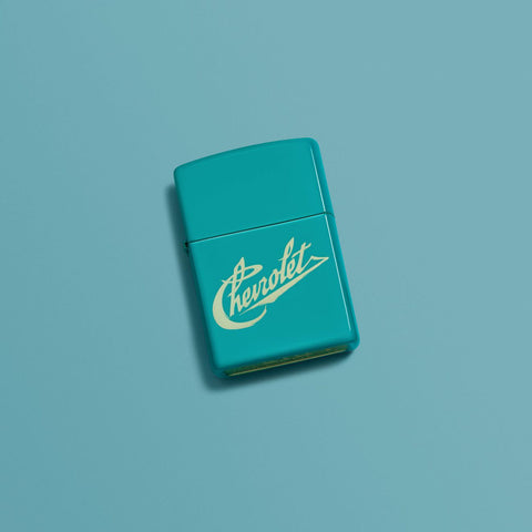 Lifestyle image of Chevy Script Logo Flat Turquoise Windproof Lighter laying on a blue turquoise background.