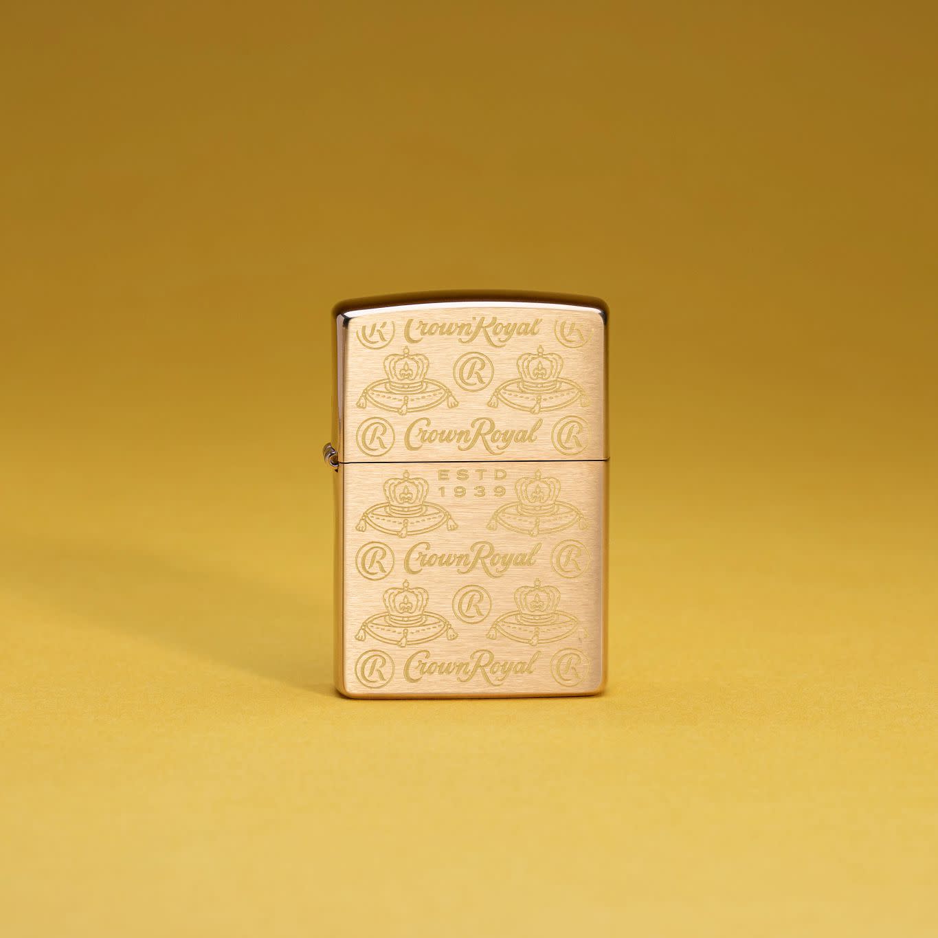 Lifestyle image of Zippo Crown Royal Elegance Design Brushed Brass Windproof Lighter standing with a mustard yellow background.