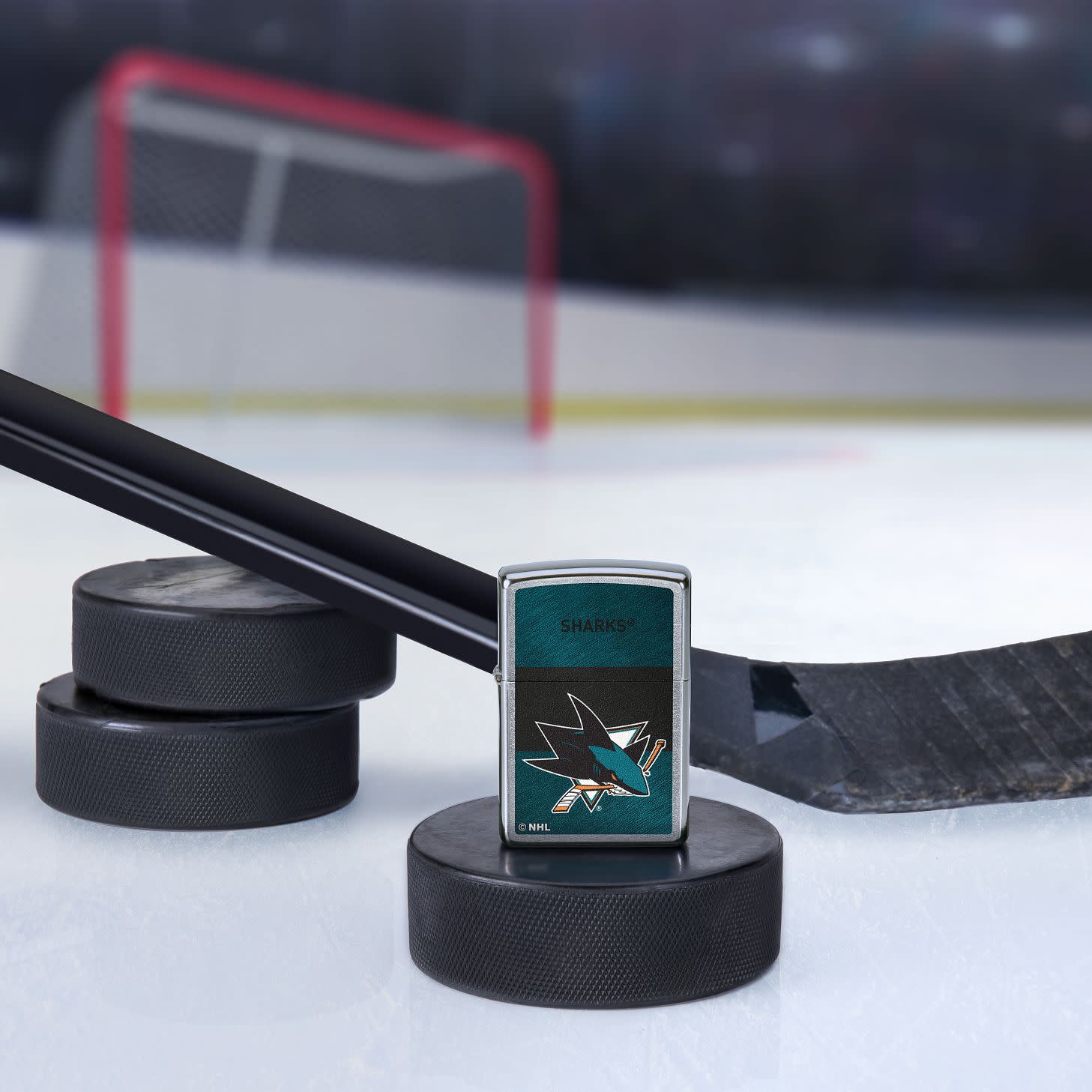 Lifestyle image of the NHL® San Jose Sharks™ Street Chrome™ Windproof Lighter standing with a hockey puck and hockey stick, with a hockey net in the background.