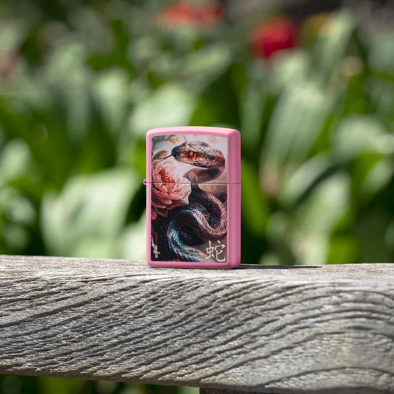 Lifestyle image of Zippo MazziÂ® Snake and Flower Design Pink Matte Windproof Lighter standing on a wooden bench with blurred flowers behind it.