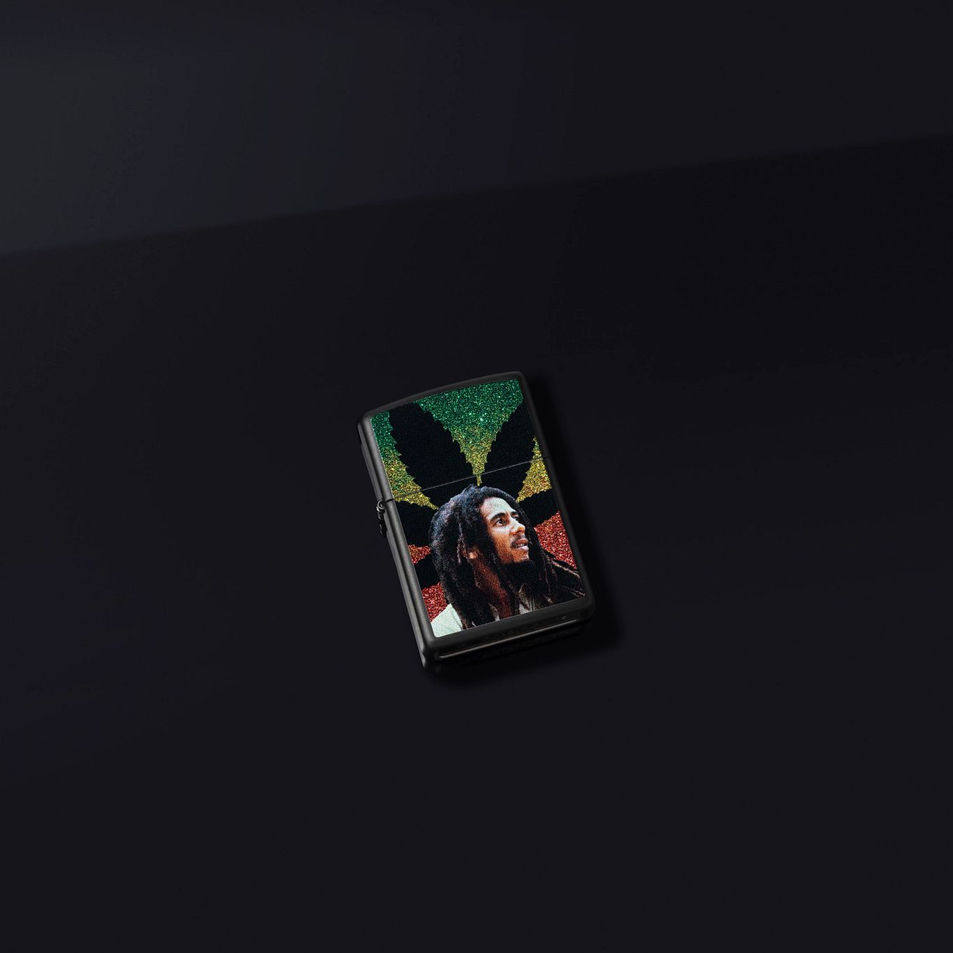Lifestyle image of Zippo Bob Marley Cannabis Design Black Matte Windproof Lighter laying on a dark gray background.