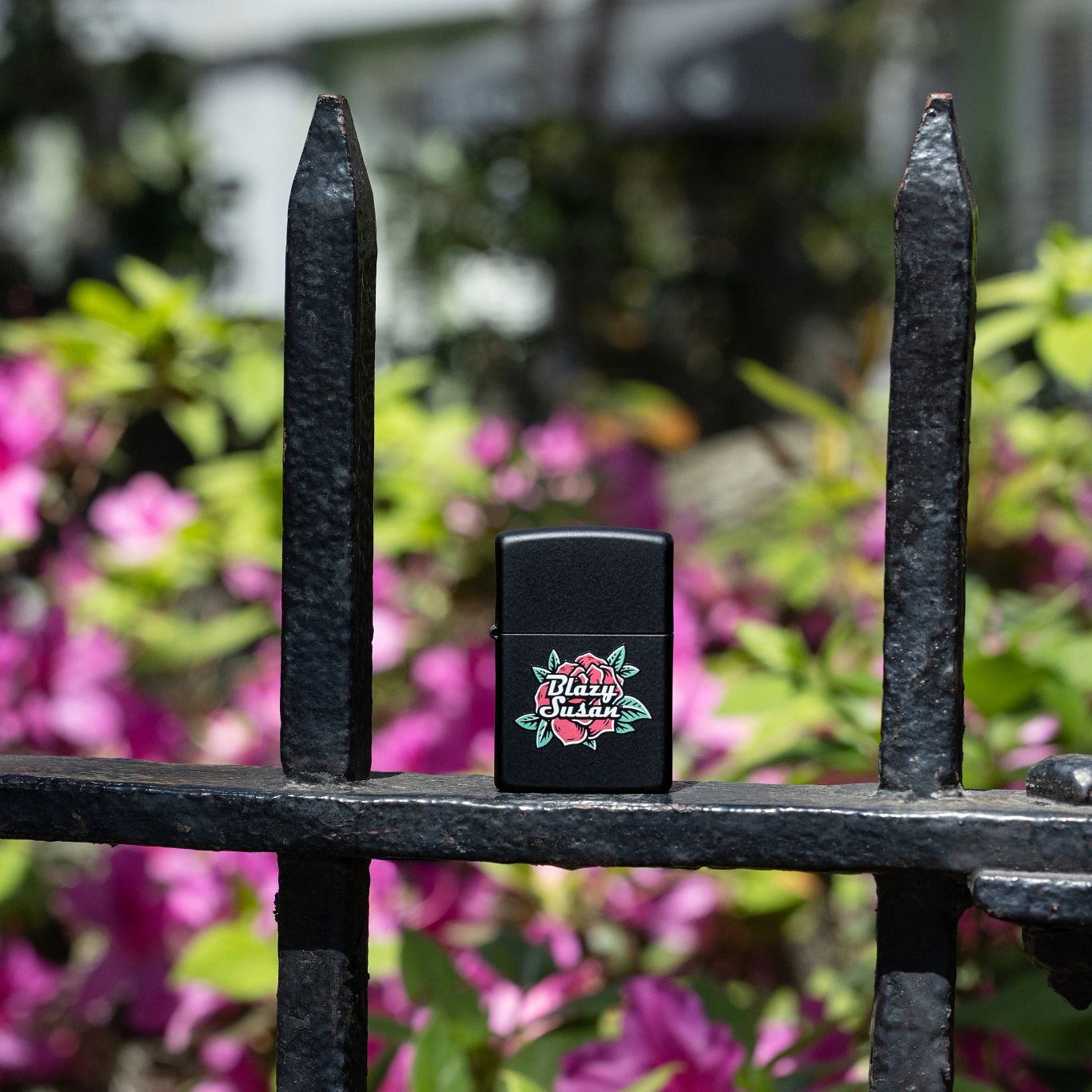 Lifestyle image of Zippo Blazy Susan™ Logo Design Black Matte Windproof Lighter standing on a wrought iron fence with blurred flowers in the background.