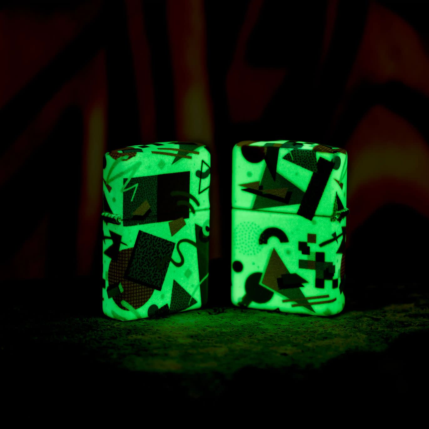 Lifestyle image of two Zippo Throwback Pattern Design 540 Color Glow in the Dark Windproof Lighters glowing in the dark, one showing the front of the lighter and the other showing the back.