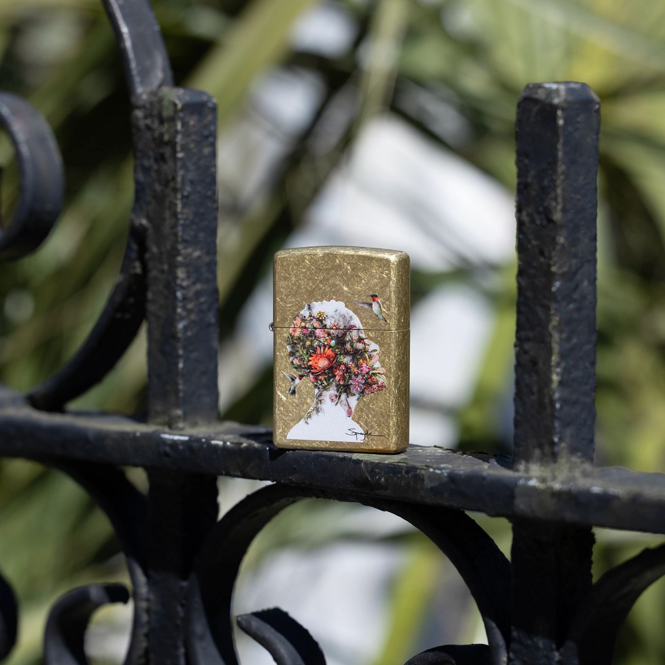 Lifestyle image of Zippo Spazuk Hummingbirds Design Street Brass Windproof Lighter standing on a wrought iron fence with blurred greenery in the background.