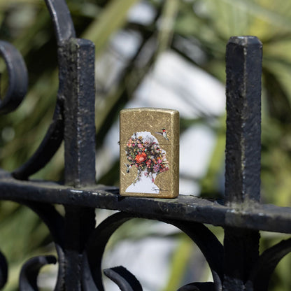 Lifestyle image of Zippo Spazuk Hummingbirds Design Street Brass Windproof Lighter standing on a wrought iron fence with blurred greenery in the background.