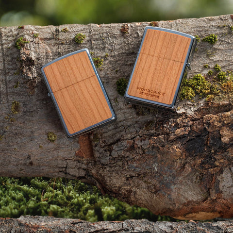 Lifestyle image of WOODCHUCK USA Cherry Emblem Windproof Lighter, laying on a log with one lighter showing the front and the other showing the back
