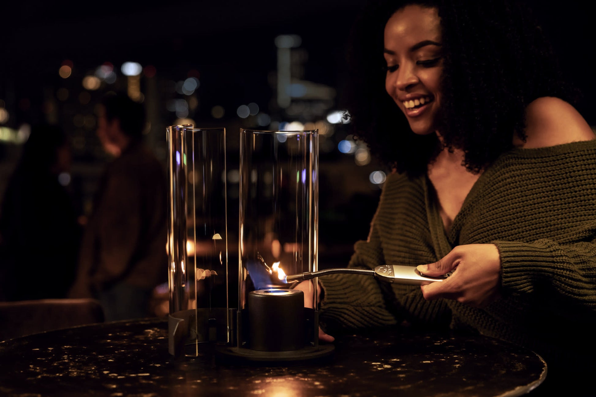 Lifestyle image of Zippo FlameScapesâ„¢ Spiral Fire Feature being lit by a woman with cityscape and couple in the background.