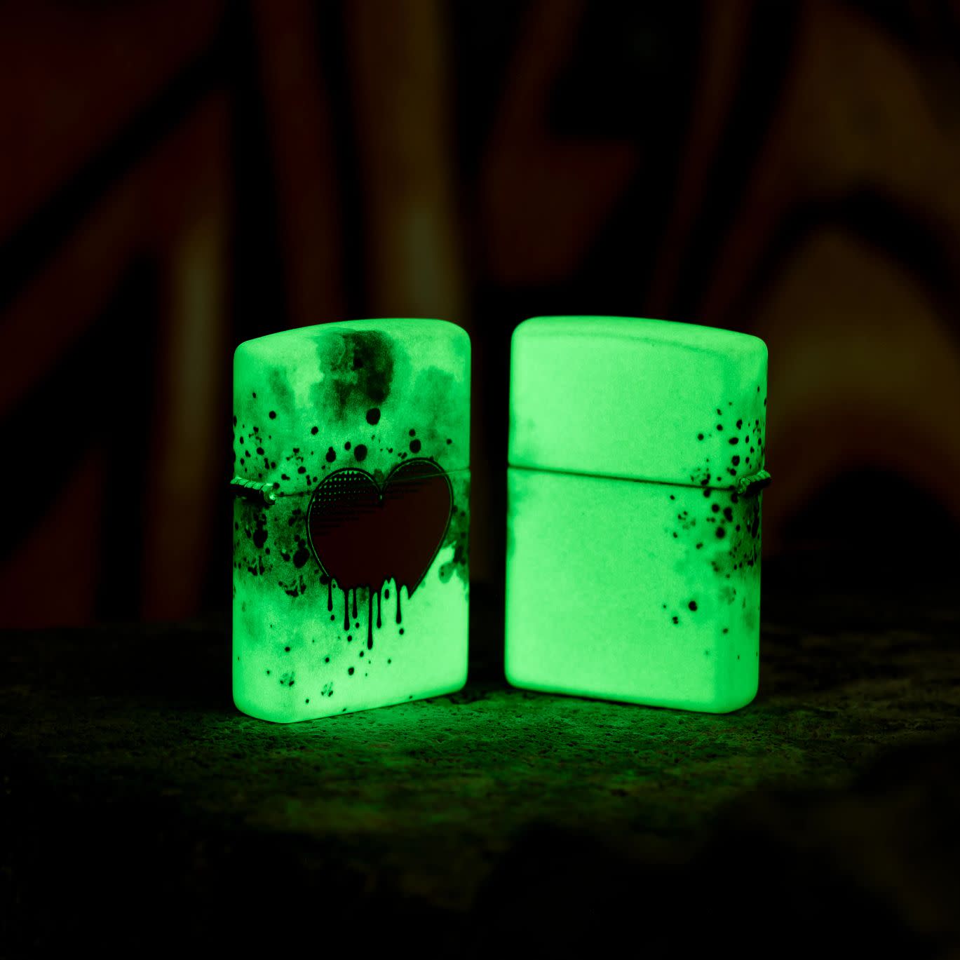 Lifestyle image of two Zippo Love Drip Design 540 Color Glow in the Dark Windproof Lighters glowing in the dark, one showing the front of the lighter and the other showing the back.
