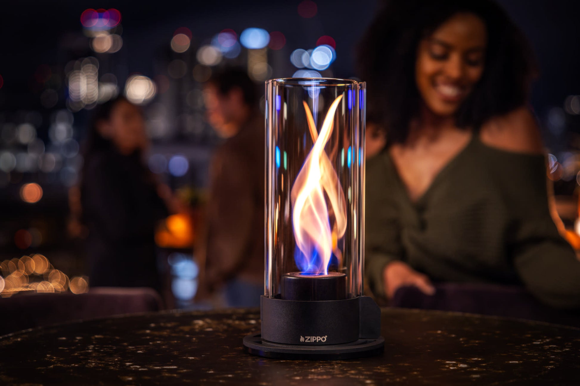 Lifestyle image of Zippo FlameScapesâ„¢ Spiral Fire Feature in black, lit on a table with a woman and couple in the background.