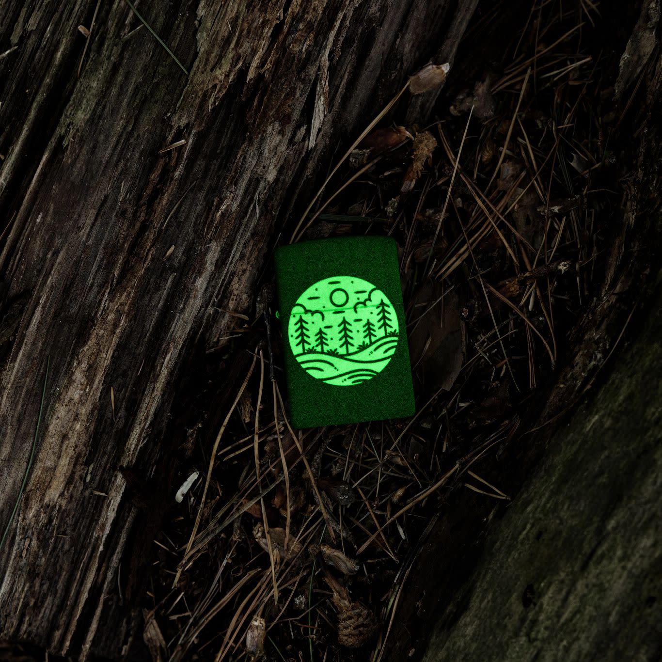 Lifestyle image of Zippo Moonlit Field Design 540 Color Glow in the Dark Windproof Lighter laying in an old log, glowing in the dark.