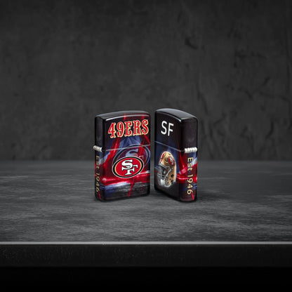 Lifestyle image of two Zippo NFL San Francisco 49ers 540 Matte Windproof Lighters, one showing the front of the lighter and the other showing the back, standing on a dark grey surface.