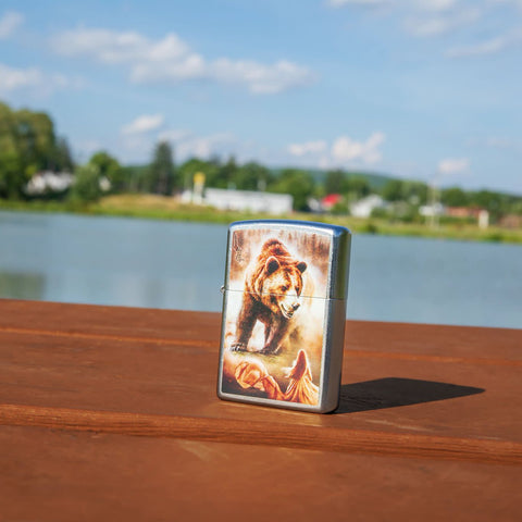 Lifestyle image of Mazzi Grizzly Bear Street Chrome Windproof Lighter standing on a railing with a lake in the background.