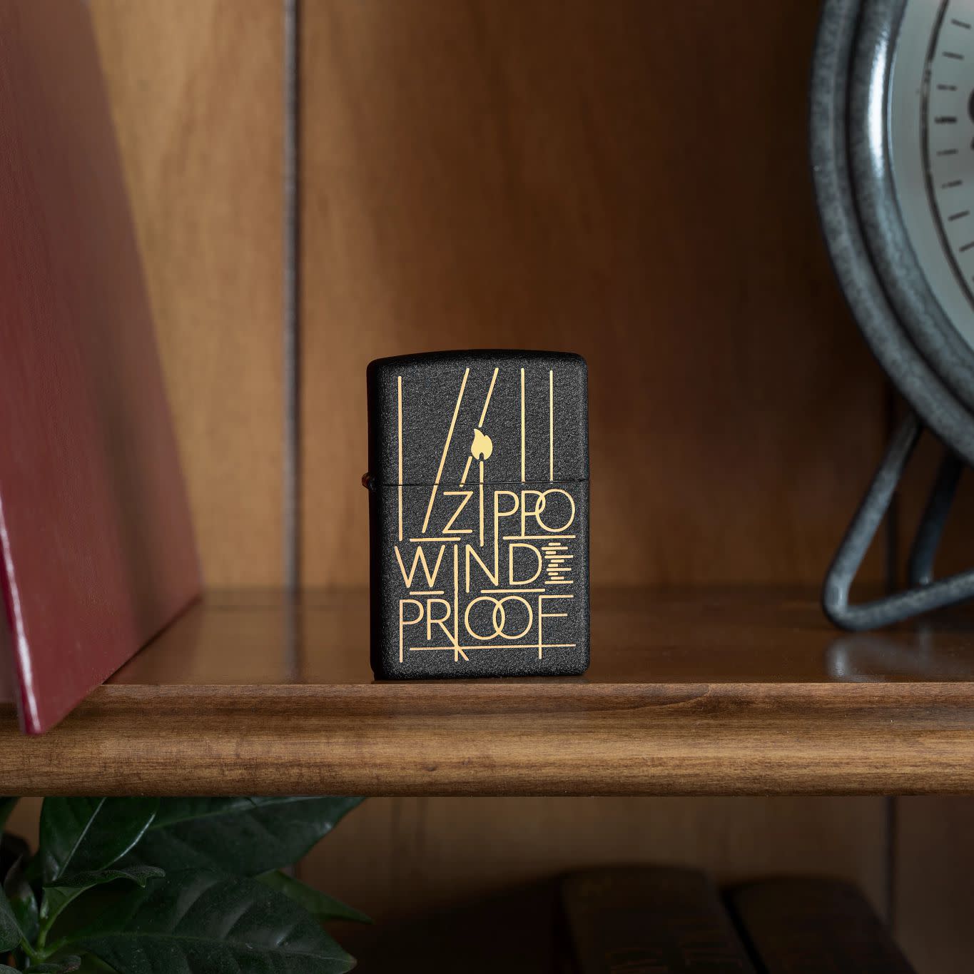 Lifestyle image of Zippo Line Art Zippo Design Black Crackle Windproof Lighter standing on a shelf beside a book and a clock.