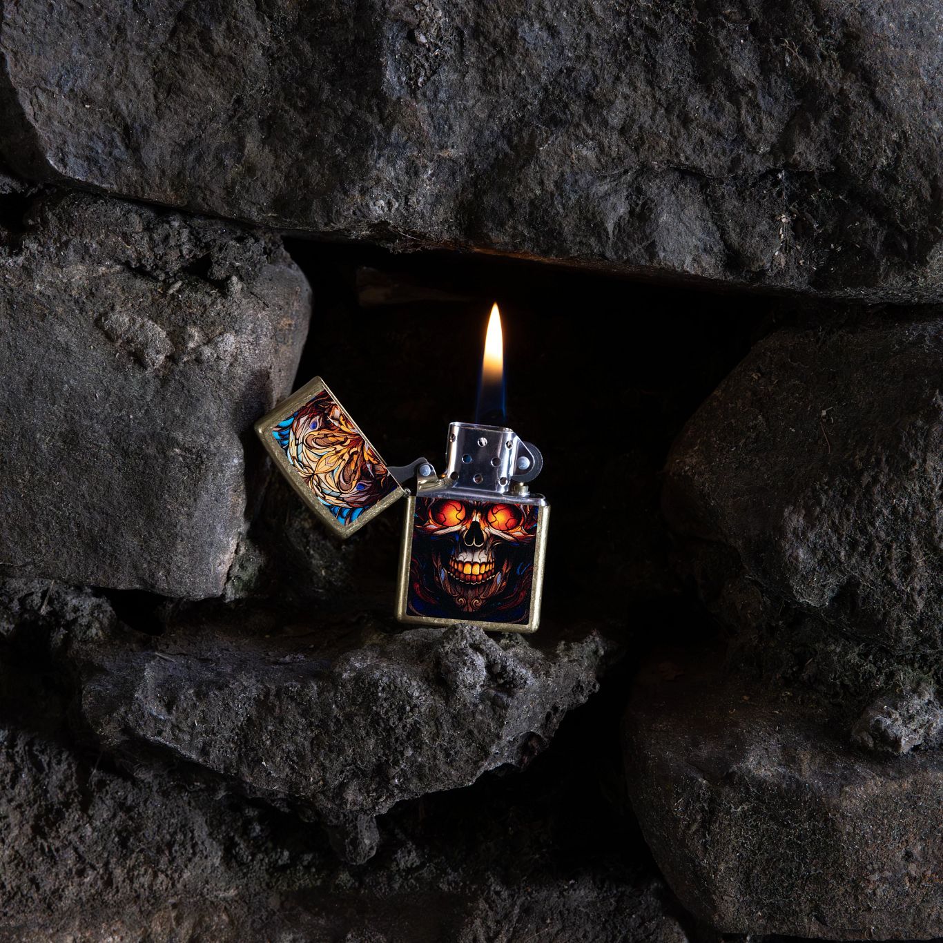 Lifestyle image of Zippo Stained Glass Skull Design Street Brass Windproof Lighter open and lit inside an opening within a stack of rocks.