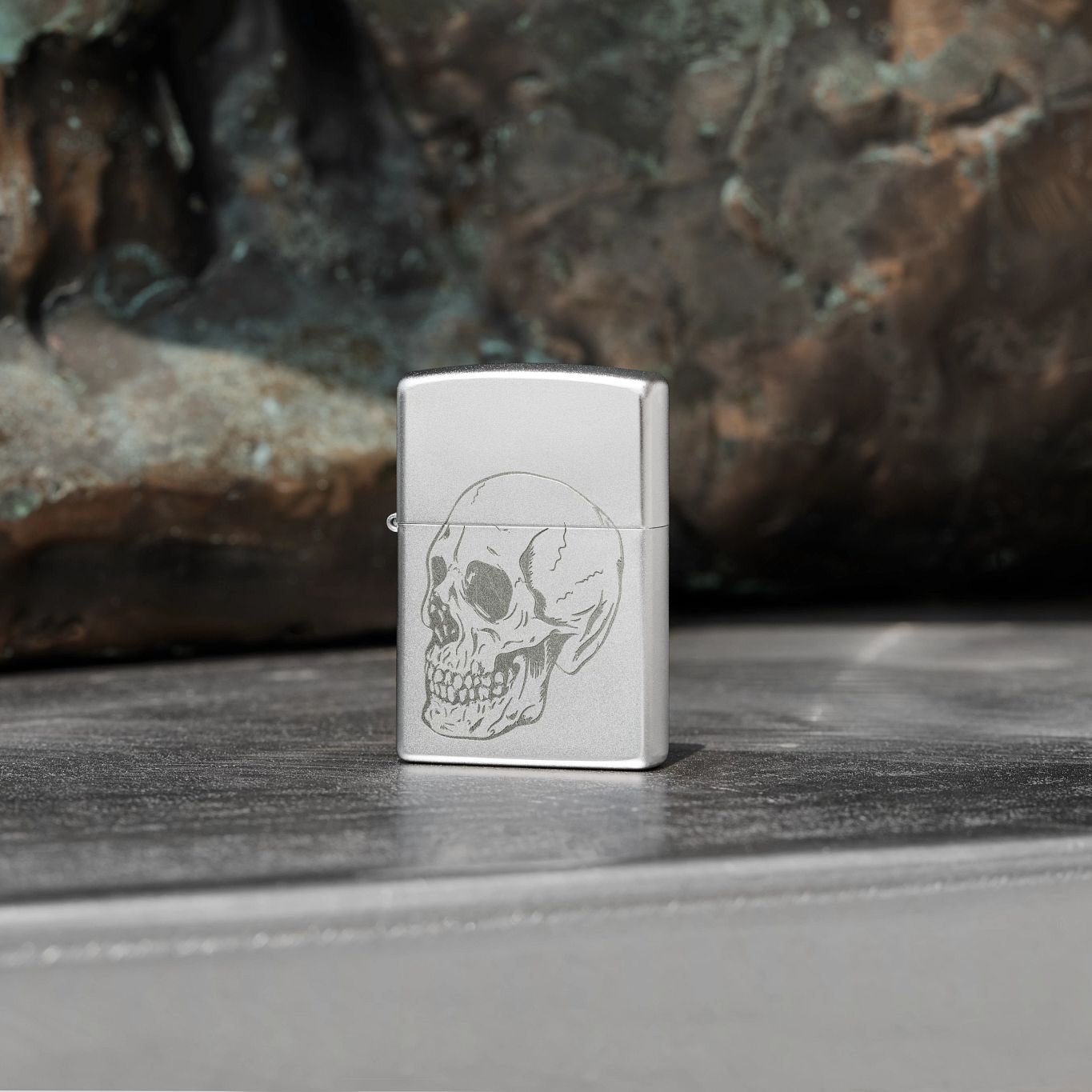 Lifestyle image of Zippo Subtle Skull Design Satin Chrome Windproof Lighter standing on a chrome surface with rock behind it.