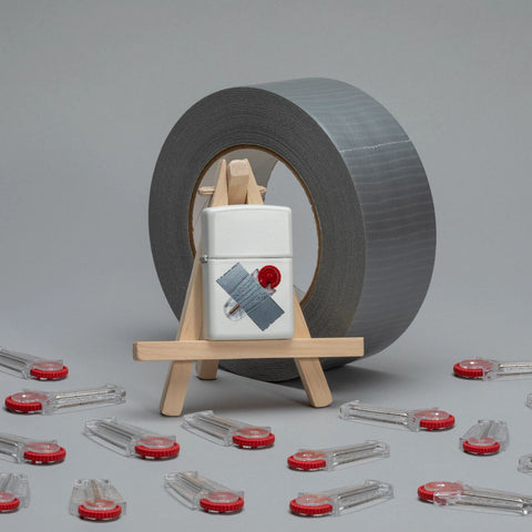Lifestyle image of Taped Flint Dispenser Design White Matte Windproof Lighter standing on an easel with a roll of duct tape behind it and flint wheel laying on the ground.