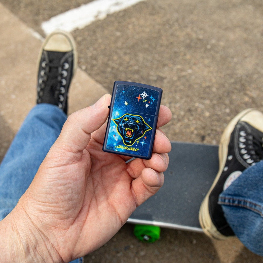 Lifestyle image of Santa Cruz Panther Navy Matte Windproof Lighter closed in hand, with a skateboard in the background