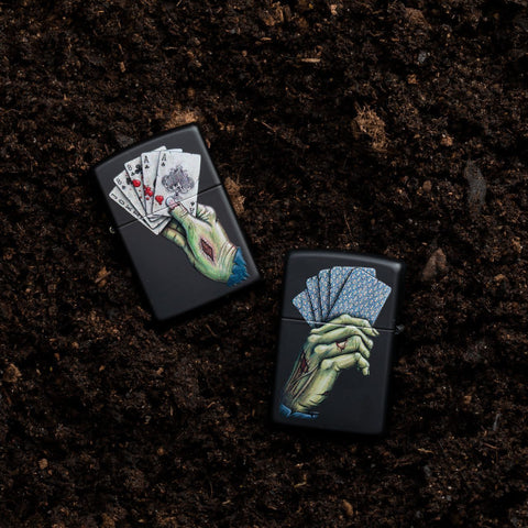 Lifestyle image of 2 Dead Mans Hand Design Black Matte Windproof Lighters, one showing the front and the other showing the back, laying in dirt