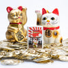 Lifestyle image of Lucky Cat Design White Matte Windproof Lighter standing with golden coins and two lucky cat statues