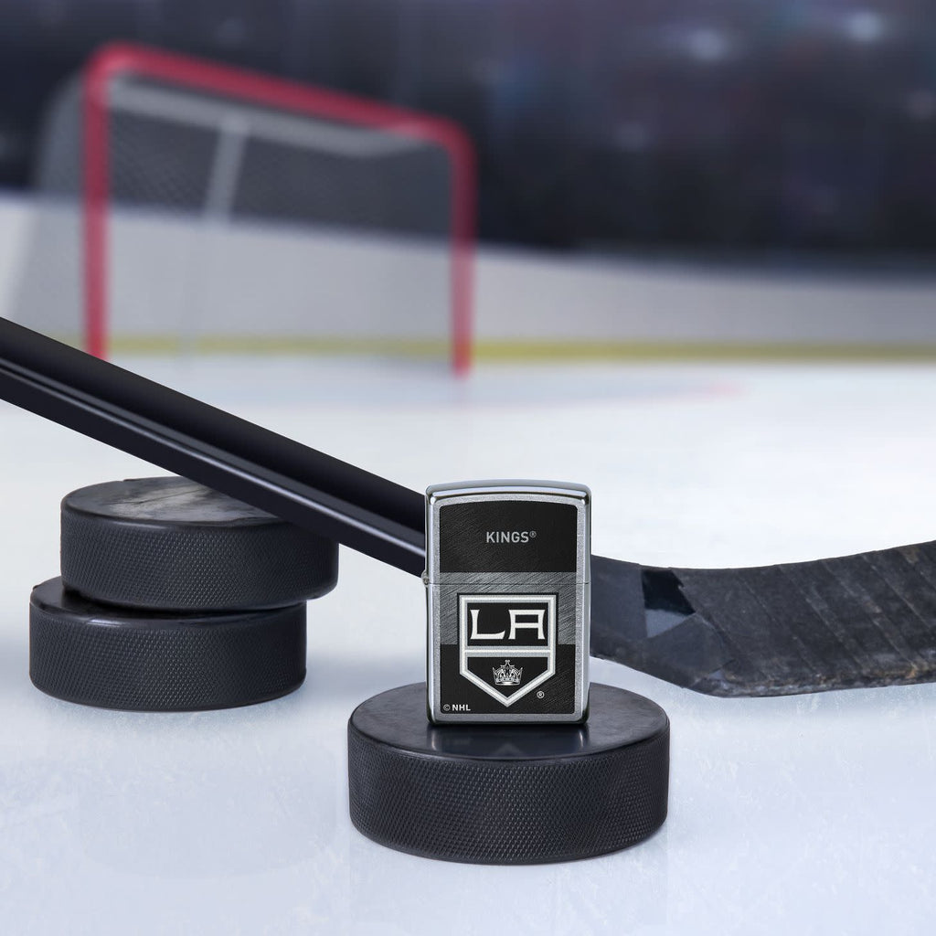 Lifestyle image of the NHL® LA Kings™ Street Chrome™ Windproof Lighter standing with a hockey puck and hockey stick, with a hockey net in the background.