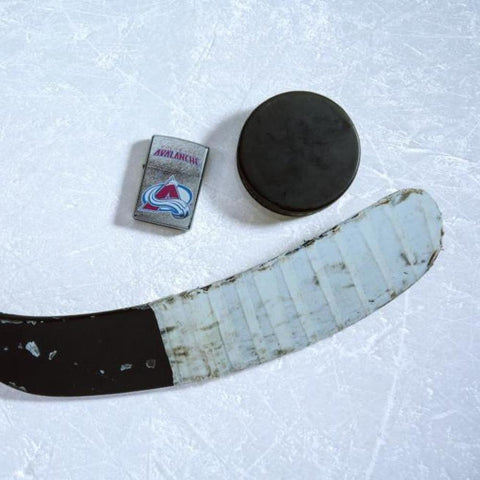Lifestyle image of the NHL Colorado Avalancher Street Chrome Windproof Lighter laying on ice with a hockey puck and stick
