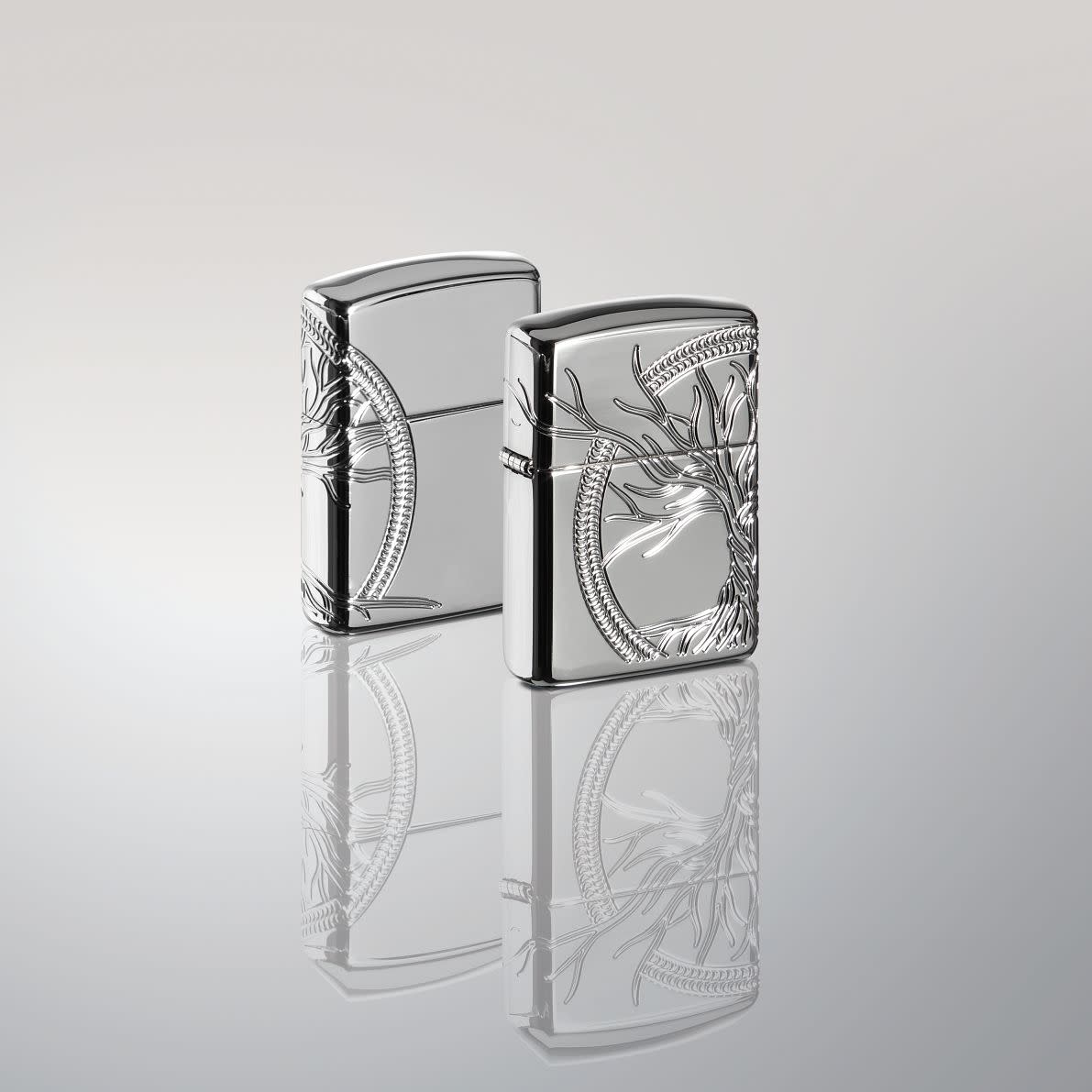 Lifestyle image of two ArmorÂ® High Polish Sterling Silver Tree of Life Windproof Lighters standing in a silver mirror scene. One lighter is showing the front of the design with the other showing the back.