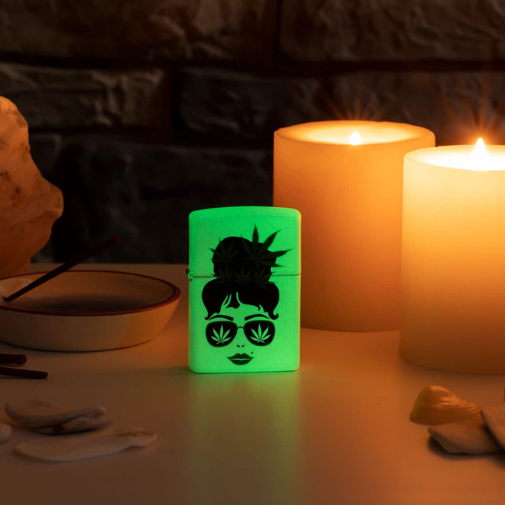 Lifestyle image of Zippo Cannabis Girl Design Glow In The Dark Pocket Lighter glowing on a side stand with candles in the background.