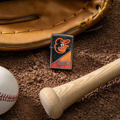 Lifestyle image of MLB™ Baltimore Orioles™ Street Chrome™ Windproof Lighter laying on a baseball field with a glove, ball, and bat.