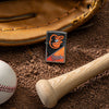 Lifestyle image of MLB® Baltimore Orioles™ Street Chrome™ Windproof Lighter laying on a baseball field with a glove, ball, and bat.