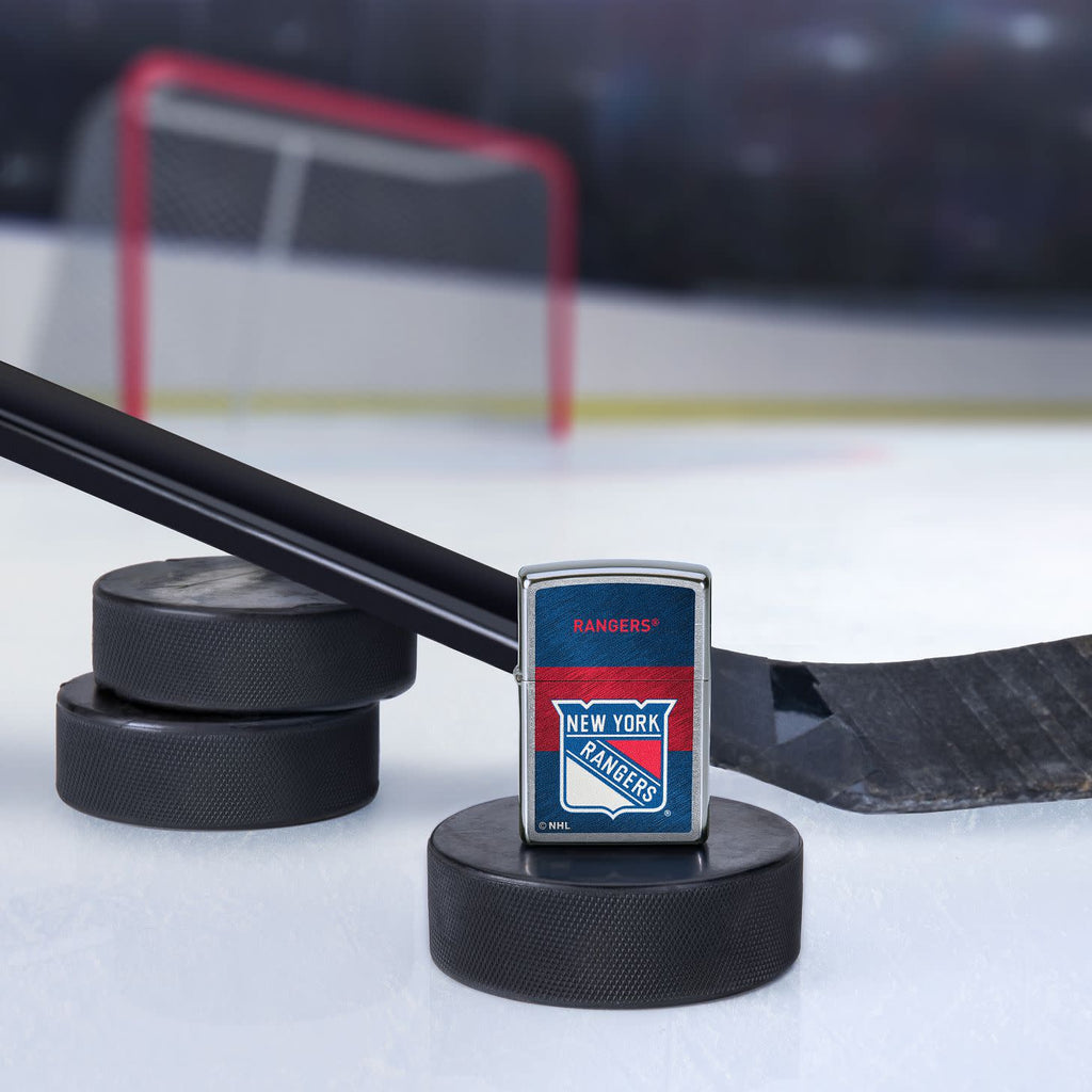Lifestyle image of the NHL® New York Rangers™ Street Chrome™ Windproof Lighter standing with a hockey puck and hockey stick, with a hockey net in the background.
