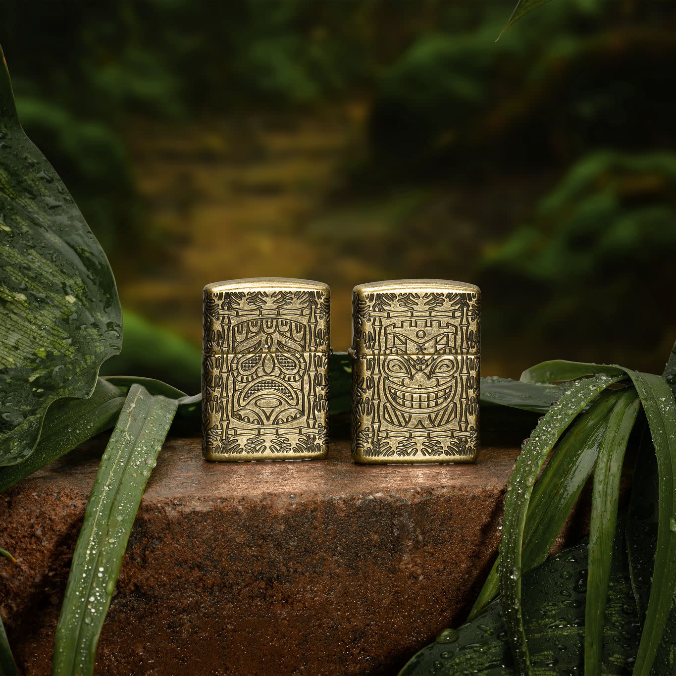 Lifestyle image of two Tiki Design ArmorÂ® Antique Brass Windproof Lighters, one showing the front of the design and the other showing the back. The lighters are standing on a stone in a jungle theme.