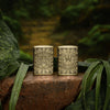 Lifestyle image of two Tiki Design Armor® Antique Brass Windproof Lighters, one showing the front of the design and the other showing the back. The lighters are standing on a stone in a jungle theme.