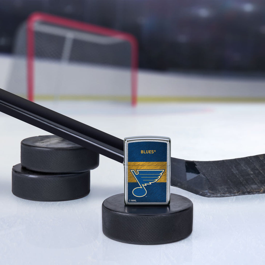 Lifestyle image of the NHL® St Louis Blues™ Street Chrome™ Windproof Lighter standing with a hockey puck and hockey stick, with a hockey net in the background.
